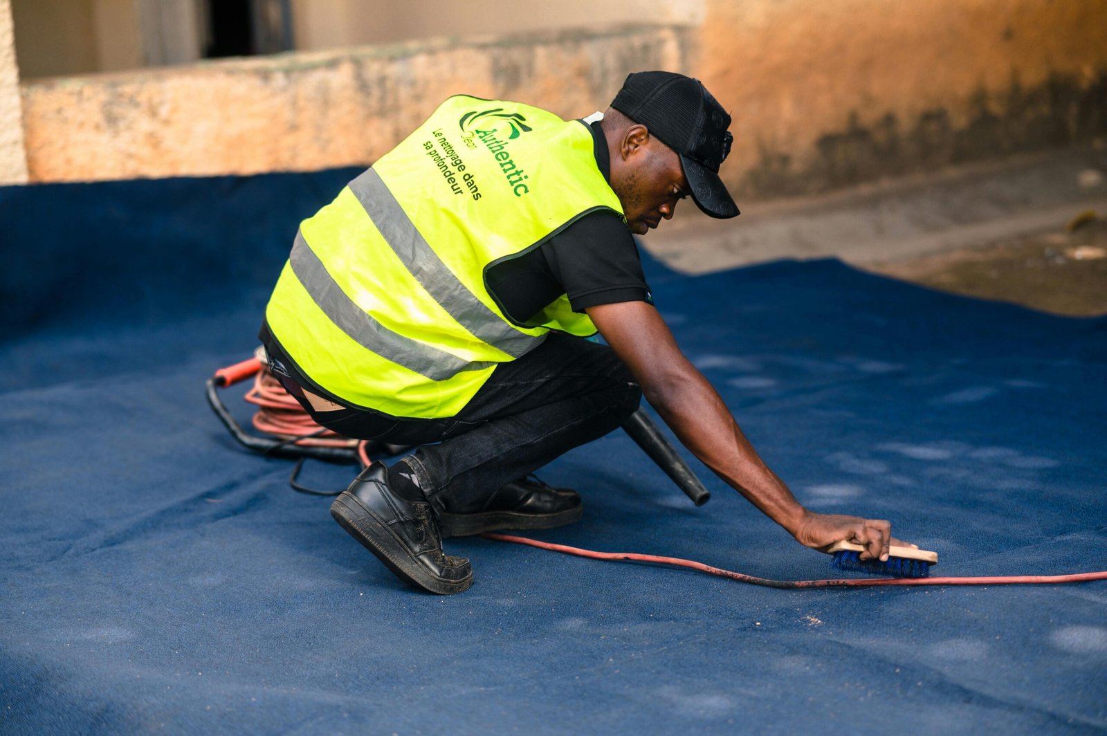 A worker dressed in a safety vest cleans a blue carpet outdoors with a brush.